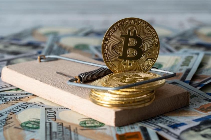 Goldene Kryptowährungsmünzen Bitcoin in einer Mausefalle, mit 100-Dollar-Scheinen im Hintergrund.